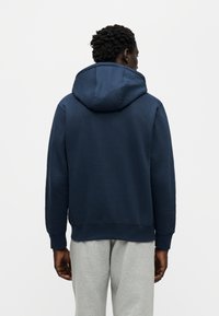 Σκούρα μπλε φούτερ με κουκούλα, κατασκευασμένο από απαλό ύφασμα, με ίσια γραμμή και ριμπ μανσέτες. Φοριέται με ανοιχτό γκρι φόρμα.