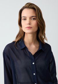 Navy blue, sheer button-up shirt with a collar, single pocket, and white buttons. Soft fabric with a relaxed fit and rolled sleeves.