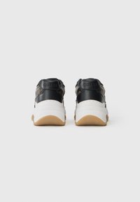 Para czarnych i brązowych sneakersów marki Guess z białymi, masywnymi podeszwami, pokazanych z tyłu na jednolitym białym tle.