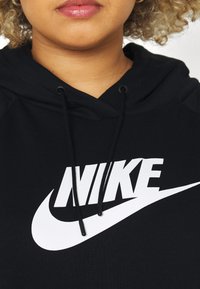 Närbild av en person som bär en svart hoodie med vit Nike-logotyp och snören, där nedre delen av ansiktet och lockigt hår syns.