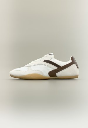 Zapatilla de ante blanco y marrón y cuero con suela de goma, cordones laterales y franja curva de acento marrón oscuro sobre fondo neutro.