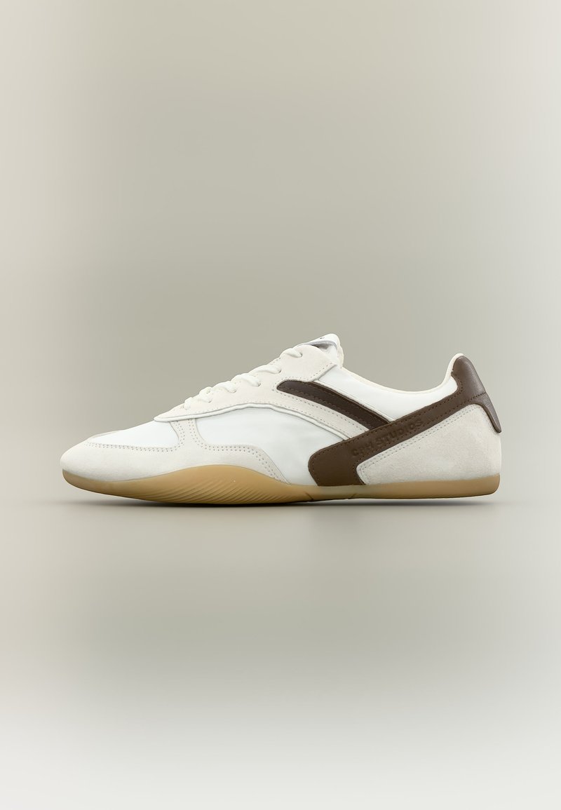 Λευκό και καφέ σουέντ και δερμάτινο αθλητικό παπούτσι με λαστιχένια σόλα, πλαϊνές κορδόνια και κυρτή σκούρα καφέ διακοσμητική λωρίδα σε ουδέτερο φόντο.