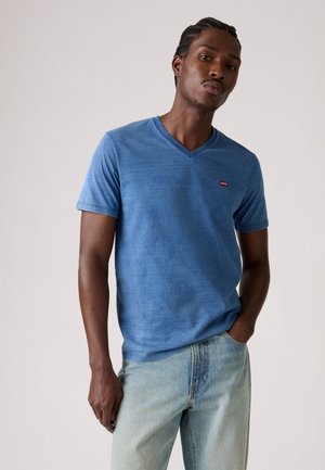 Blaue V-Ausschnitt-T-Shirt aus weichem Stoff, mit einem kleinen roten Logo auf der Brust. Getragen mit hellblauen Jeans.