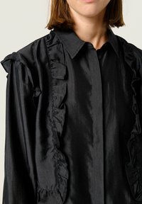 Black blouse featuring a satin finish, front button closure, and contrasting ruffled trim along the placket and shoulders.