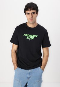 Juodas medvilninis marškinėliai su žaliais užrašais "ORDINARY ELITE" ir logotipu, derinami su šviesiai mėlynomis džinsinėmis kelnėmis, kurių kišenėse yra detalės.
