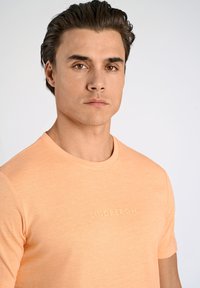 T-shirt a maniche corte, con scollo rotondo, di colore arancione chiaro, realizzato in tessuto morbido, con un logo "Lindbergh" in rilievo sobrio sul petto.