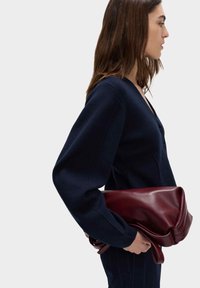 Granatowy sweter o dzianinowym splocie z głębokim dekoltem w kształcie litery V i długimi rękawami, zestawiony z bordową skórzaną torebką, o gładkiej teksturze i luźnym fasonie.