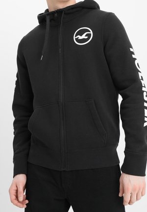 Henkilö, joka pukeutunut mustaan vetoketjulliseen huppariin, jossa valkoinen lokkikuvio rinnassa ja "HOLLISTER"-teksti oikeassa hihassa.