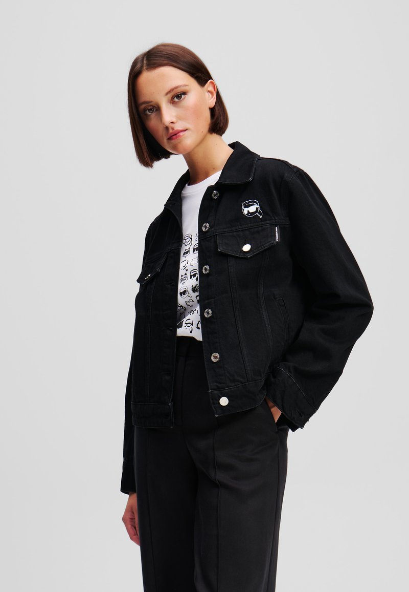 Črna denim jakna se ponaša s klasičnim dizajnom, dvema žepoma na prsih, srebrnim okovjem in izvezenim logotipom na levi strani.