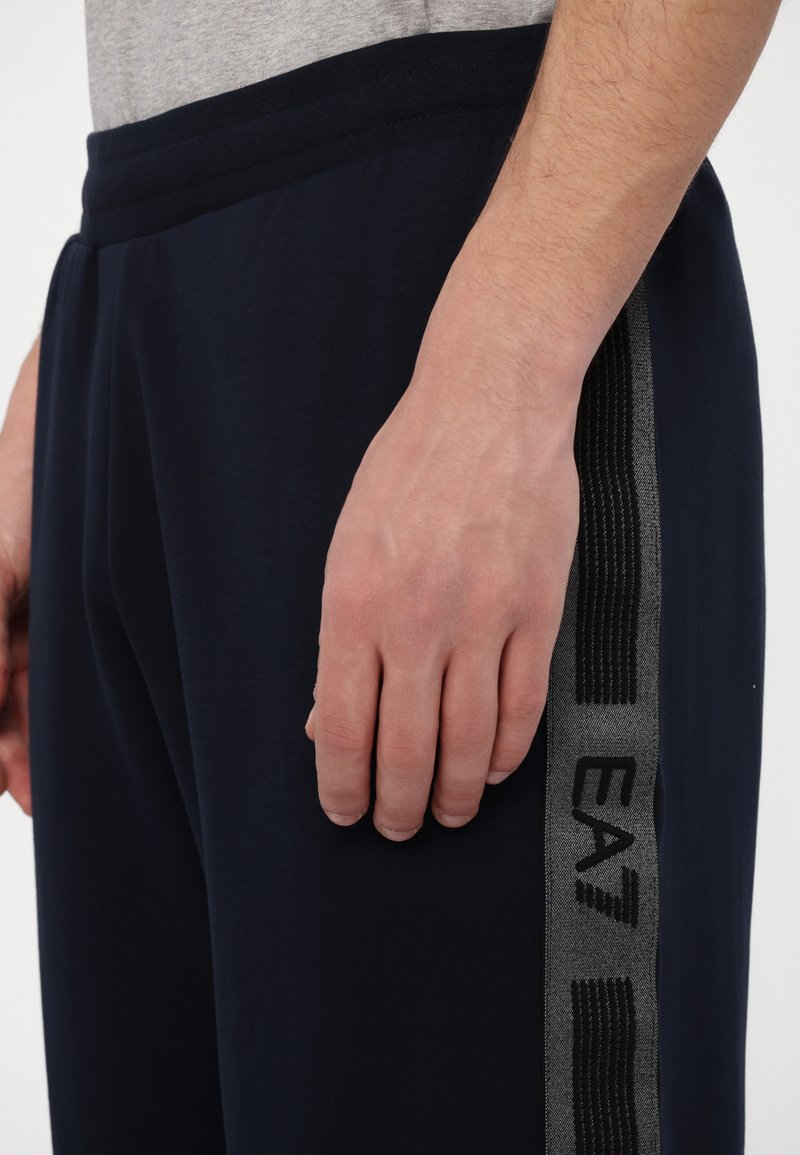 Joggers blu navy con vita regolabile, dotati di una striscia laterale grigia con dettagli testurizzati e un logo stampato. Tessuto morbido e liscio.