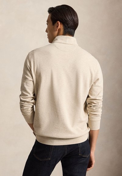 Man die een beige lange-mouwen trui en donkerblauwe spijkerbroek draagt, staat met zijn handen in zijn zakken, met zijn rug naar een effen beige achtergrond.