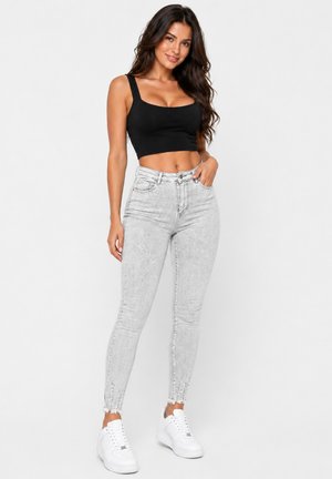 Frau mit langen dunklen Haaren, die ein schwarzes Croptop, hellgraue hoch taillierte Skinny Jeans und weiße Sneakers trägt und mit einer Hand in der Tasche steht.