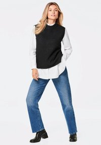 Zwarte gebreide mouwloze top over een witte blouse met kraag, gecombineerd met blauwe recht gesneden jeans en zwarte block-heeled enkellaarsjes.