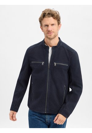 Mann trägt marineblaue Jacke mit Reißverschluss und zwei Brust- und zwei seitlichen Reißverschlusstaschen, steht vor weißem Hintergrund.