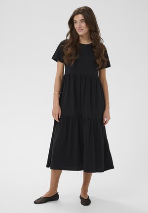 Junge Frau mit langen braunen Haaren, die ein schwarzes, kurzärmliges, gestuftes Midikleid und schwarze Netz-Ballerinas trägt, stehend vor einem schlichten Hintergrund.