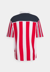 Polo tričko s krátkým rukávem, které má červené a bílé svislé pruhy s pevným tmavě modrým zadním dílem a límcem. Vyrobeno z bavlněné směsi.