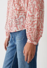 Blusa leggera a maniche lunghe in crema con motivo a foglie rosse; presenta polsini arricciati e un taglio dritto. Abbinata a jeans in denim blu.