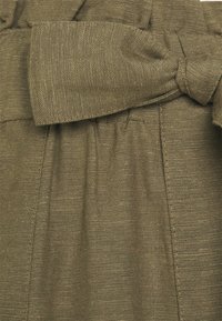 Gros plan sur un tissu texturé vert olive rassemblé à une couture avec une large ceinture nouée dans le même matériau.