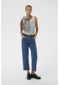 Top a righe, maglione grigio annodato attorno al collo, jeans in denim blu, cintura nera e mocassini neri. Modella in piedi con le mani nelle tasche.