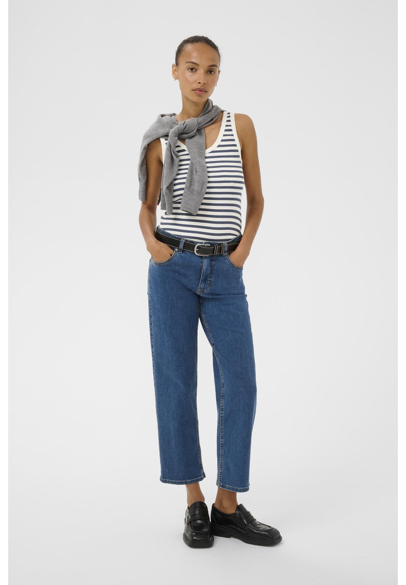 Top a righe, maglione grigio annodato attorno al collo, jeans in denim blu, cintura nera e mocassini neri. Modella in piedi con le mani nelle tasche.