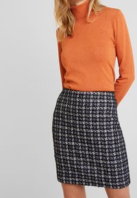 Pomarańczowy sweter z kołnierzem typu golf, zestawiony z granatową spódnicą o fakturowanej powierzchni, zdobioną białym geometrycznym wzorem. Stylizacja ma dopasowany fason.