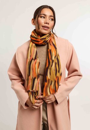 Sciarpa multicolore con strisce astratte arancioni, gialle e marroni. Tessuto leggero con bordi sfrangiati, indossata sopra una maglietta beige.