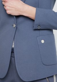 Osoba zapięta niebieską marynarkę z srebrnymi guzikami, eksponująca część dopasowanych niebieskich spodni z detalem zamka błyskawicznego.