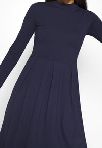 Granatowa sukienka z długim rękawem ma wysoki dekolt i plisowany spódnicę, wykonana z gładkiego, elastycznego materiału, który elegancko układa się na sylwetce.