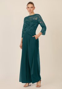 Tuta teal con corpino in pizzo, maniche a tre quarti e pantaloni larghi pieghettati. Presenta una tasca laterale e una morbida texture in satin.