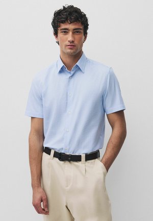 Mann trägt hellblaues, kurzärmliges Hemd mit Knöpfen, eingesteckt in beige Hose mit schwarzem Gürtel, steht vor schlichtem hellem Hintergrund.