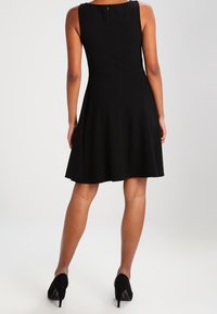 Kvinna som bär en ärmlös, knälång svart klänning och svarta högklackade skor, står med ryggen mot kameran på en vit bakgrund.