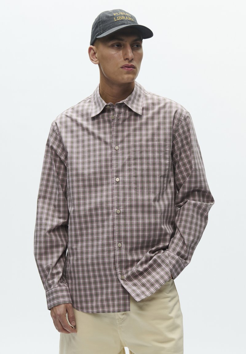 Camisa de manga larga a cuadros rojos y verdes con cuello abierto y cierre de botones. Se lleva con pantalones beige y una gorra negra.