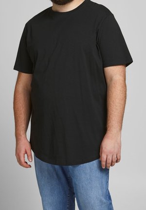Homme portant un t-shirt noir uni à manches courtes et un jean bleu sur un fond gris neutre.
