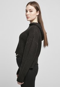 Fekete, rövidített kapucnis pulóver, texturált kötött mintával, oversized ujjakra és kapucnira, magas derekú fekete farmernadrággal viselve.