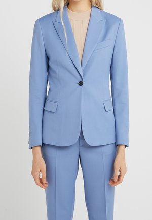 Frau trägt einen hellblauen, maßgeschneiderten Anzug mit einem Ein-Knopf-Blazer und passenden Hosen über einem beigen, gerippten Top, steht vor einem schlichten Hintergrund.
