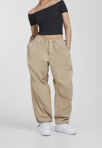 Beige, loose-fitting cargo pants with an elastic waistband and side pockets, paired with a black off-the-shoulder top and white sneakers.