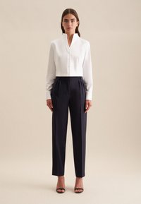 Witte blouse met hoge kraag en lange mouwen, gecombineerd met marineblauwe broek. De broek heeft plooien en een getailleerde pasvorm. Hakken accentueren de look.