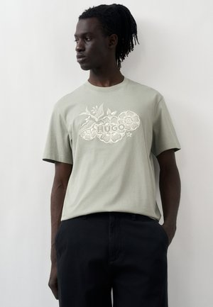 Mężczyzna z dredami w jasnozielonej koszulce HUGO z motywem kwiatowym i ptaków, w połączeniu z czarnymi spodniami, stojący przy białej ścianie.