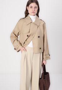 Cappotto trench cropped beige con ampi revers, bottoni doppi e spalline. Abbinato a pantaloni color beige chiaro e una borsa marrone.