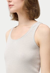 Närbild av en kvinna med axellångt brunt hår som bär en ljusbeige, ribbad ärmlös topp mot en enfärgad bakgrund.