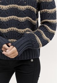 Marine gebreide trui met crèmekleurig zigzagpatroon, voorzien van geribbelde manchetten en zoom, ruime mouwen en open gebreide details. Gemaakt over zwarte broek.