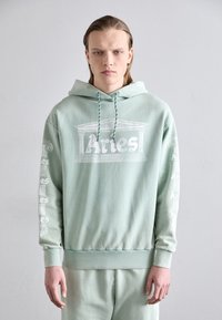 Hoddie vert menthe en tissu doux. Présente un logo et des graffitis à l'avant et sur les manches. Cordon de serrage sur la capuche et poignets côtelés.