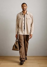 Bărbat care merge pe interior, purtând o cămașă bej cu nasturi și pantaloni maro, având în mâna stângă o geantă tote maro asortată.