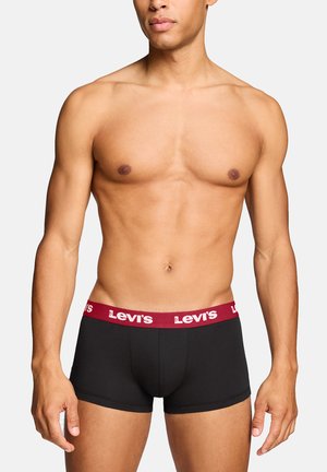 Männlicher Torso, der schwarze Levi's Boxershorts mit rotem Bund trägt, auf dem das Levi's-Logo zu sehen ist, vor einem einfarbigen hellen Hintergrund.