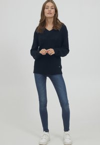 Maglione lavorato a maglia di un blu navy scuro con scollatura a V, polsini a costine e un dettaglio con logo laterale, abbinato a jeans slim-fit e sneakers bianche.