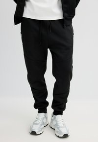 Pantalones jogger negros con acabado texturizado, con bolsillos laterales, cintura elástica y diseño de pierna cónica, combinados con zapatillas blancas.