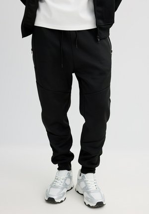 Pantalones jogger negros con acabado texturizado, con bolsillos laterales, cintura elástica y diseño de pierna cónica, combinados con zapatillas blancas.