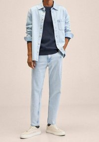 Camisa de mezclilla azul claro sobre un suéter azul marino, combinada con jeans azul claro y zapatillas crema. Cuenta con un bolsillo en el pecho y botones en la parte frontal.