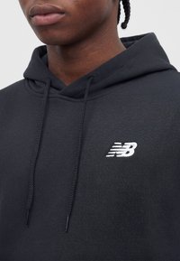 Nærbilde av en person som har på seg en svart hettegenser med snorer og en liten hvit New Balance-logo på brystet.