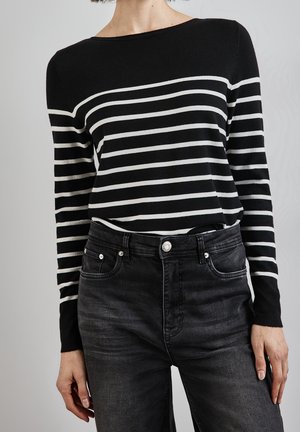 Femme portant un haut à manches longues rayé noir et blanc rentré dans un jean noir délavé taille haute, debout devant un fond uni.